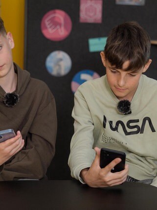Zwei Schüler sitzen in einem modernen Klassenzimmer. Beide schauen genervt auf ihr Smartphone.