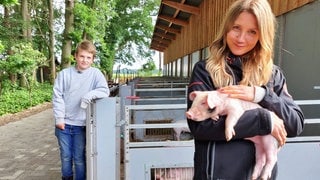 neuneinhalb-Reporterin Jana und der 9-jährige Hinnerk stehen vor den Schweineställen. Jana hält ein kleines Ferkel im Arm.