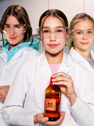 Im Hintergrund ein Labor, im Vordergrund drei Mädchen in Laborjacken und Schutzbrillen, die wissenschaftliche Instrumente halten
