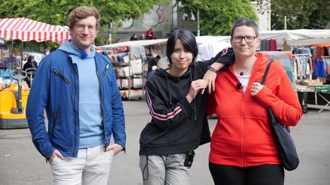 neuneinhalb-Reporter Robert steht mit dem 14-jährigen Alex und seiner Mutter Mel auf einem Wochenmarkt. Alex lehnt sich mit einem Arm auf die Schulter seiner Mutter.